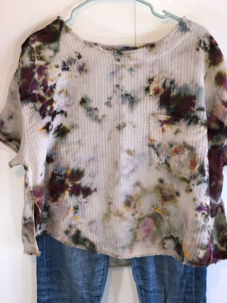 May include: A tie-dye short-sleeve shirt with a pocket, displayed on a hanger. The shirt features a ribbed texture and a mix of colors, including purple, green, and yellow, against a white background. Frayed edges add a casual touch. Paired with blue jeans.