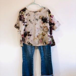 May include: A tie-dye short-sleeve shirt with a pocket and blue jeans with rolled-up cuffs hang on a hanger against a white wall. The shirt has a white base with purple, green, and brown accents. The jeans are a medium wash.
