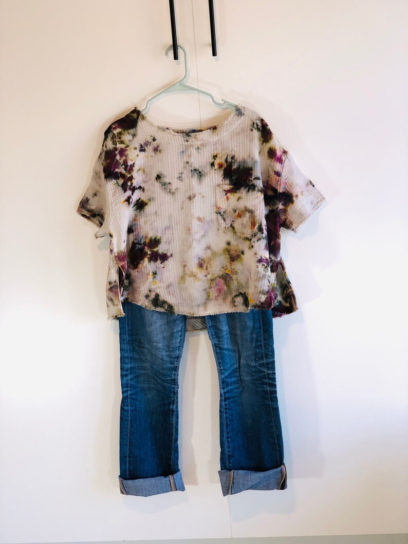 May include: A tie-dye short-sleeve shirt with a pocket and blue jeans with rolled-up cuffs hang on a hanger against a white wall. The shirt has a white base with purple, green, and brown accents. The jeans are a medium wash.