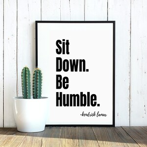 May include: Black and white framed print with the quote "Sit Down. Be Humble." attributed to Kendrick Lamar. The print is displayed on a white wood wall with a potted cactus in front of it.