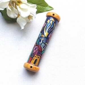 May include: A hand-painted mezuzah case with a cylindrical shape. The case features a vibrant design of colorful buildings and abstract shapes on a blue background, with wooden end caps. The artwork includes gold accents.