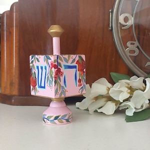May include: A pink wooden dreidel featuring hand-painted pomegranate branches with red fruit and green leaves. The Hebrew letters Shin and Hey are painted in blue on the front faces. The dreidel has a gold-painted top and a pink base with leaf details.