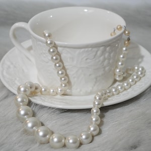 May include: A cream-coloured pearl necklace rests on a white teacup and saucer. The teacup and saucer feature a raised leaf design. The pearls are of varying sizes, with the largest at the necklace's base. The background has a soft, white, furry texture.