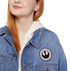 Può includere: Giacca di jeans blu con una toppa rotonda con un simbolo nero e le parole "REBELLIONS ARE BUILT ON HOPE NO". La giacca è indossata sopra una felpa con cappuccio color crema.