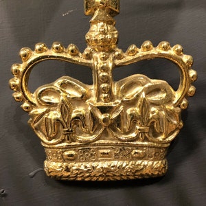 Könnte beinhalten: Eine goldfarbene Krone mit einem Kreuz oben. Die Krone hat ein detailliertes Design mit Fleur-de-lis und einem Perlenrand.