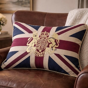 May include: Rectangular decorative pillow featuring the Union Jack flag design in navy blue, burgundy, and beige. The pillow has a gold embroidered emblem and a gold rope trim. It rests on a brown leather armchair.