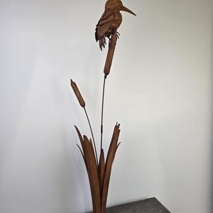 Op de afbeelding: Een roestige metalen sculptuur van een vogel die op lisdodden zit. De vogel heeft gedetailleerde veren en een lange snavel. De lisdodden en de basis zijn ook van metaal, waardoor een samenhangend, door de natuur geïnspireerd kunstwerk ontstaat.