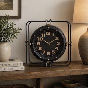 Könnte beinhalten: Eine schwarze analoge Uhr mit goldfarbenen Ziffern und Zeigern, eingefasst in einem quadratischen schwarzen Metallrahmen. Die Uhr steht auf einer Holzoberfläche, mit einer Lampe und einer Vase im Hintergrund. Das Zifferblatt hat ein klassisches, Vintage-Design.