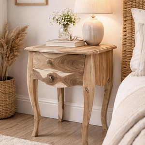 Op de afbeelding: Een houten nachtkastje met een enkele lade en gebogen poten. Bovenop staat een lamp met een beige kap, naast een vaas met witte bloemen en een stapel boeken. Het nachtkastje is van lichtgekleurd hout.