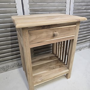 May include: A light-colored wooden nightstand with a single drawer and a lower shelf. The nightstand has a rectangular top with rounded edges and vertical slats on the sides. The drawer has a round wooden knob.