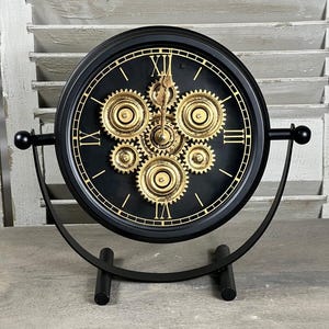 Peut inclure: Horloge de bureau de style steampunk noir et or. L'horloge présente un cadran noir avec des chiffres romains dorés et des engrenages visibles. Elle est soutenue par un support en métal noir avec un design incurvé et des boules.