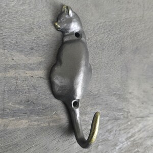 Op de afbeelding: Een gietijzeren katvormige wandhaak in een zittende pose. De haak is donkergrijs geschilderd met gouden accenten op de oren, neus en haakpunt. De haak is ongeveer 15 cm lang.