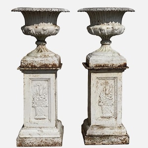 Puede incluir: Dos antiguas urnas de hierro fundido blanco con detalles ornamentados. Cada urna presenta un cuenco acanalado, un pedestal decorativo y una base cuadrada. Los pedestales tienen un panel rectangular con un diseño floral. Las urnas muestran signos de edad y desgaste.