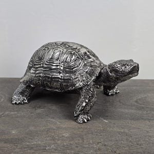 May include: A silver-colored turtle figurine with a textured shell and detailed legs. The decorative sculpture is made of metal and sits on a wooden surface. The turtle is a decorative item.