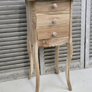 May include: A light-coloured wooden bedside table with three drawers and a small top. The bedside table has curved legs and decorative floral carvings on the side. The knobs on the drawers are round and made of wood.