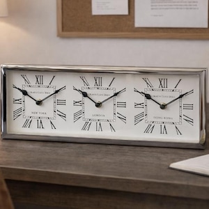 Puede incluir: Un reloj rectangular con marco plateado y tres esferas que muestran las zonas horarias de Nueva York, Londres y Hong Kong. Las esferas blancas tienen números romanos y manecillas negras. El reloj está sobre un escritorio de madera.