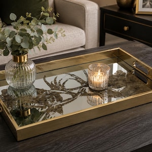 May include: A rectangular serving tray with a polished gold metal frame and a mirrored base featuring a decorative stag head design. The tray includes integrated side handles and holds a clear ribbed glass vase with greenery and a lit candle.