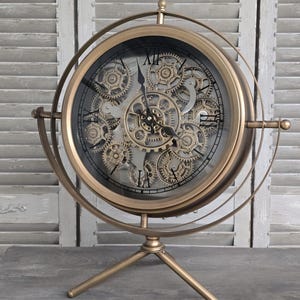Könnte beinhalten: Eine goldfarbene mechanische Uhr mit sichtbaren Zahnrädern und römischen Ziffern. Die Uhr steht auf einem Dreibeinständer und ist in einem kreisförmigen Rahmen eingefasst. Der Hintergrund zeigt eine graue Holzoberfläche und ein Fenster mit Fensterläden.