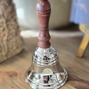Puede incluir: Una campana de plata con un mango de madera. La campana tiene un diseño decorativo alrededor del borde y las palabras "It's" y "Time" están grabadas en el lateral.
