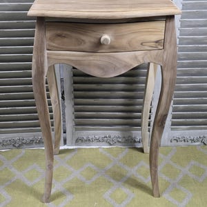 May include: A light-coloured wooden bedside table with a single drawer and curved legs. The drawer has a round wooden knob. The table has a small top and is approximately 61 cm tall.