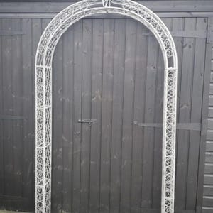 May include: A white metal garden arch with a decorative fleur-de-lis at the top. The arch features an arched top and ornate scrollwork design. The structure is freestanding and suitable for outdoor use, such as a garden or pathway.
