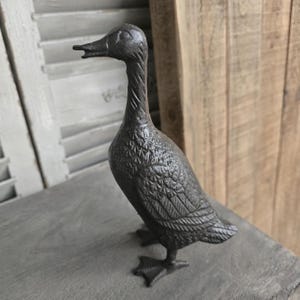 May include: A dark grey cast iron duck ornament with detailed feather and wing textures. The duck has an open beak and is standing on a grey surface. The duck is a decorative item.