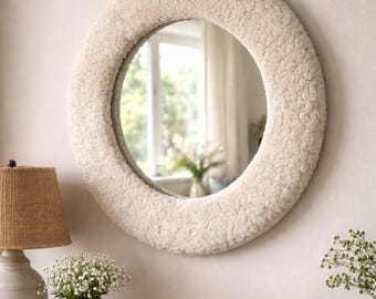 May include: A circular wall mirror featuring a thick, textured, off-white faux shearling frame. The mirror surface is reflective and round, showing a blurred interior reflection of a window and greenery.