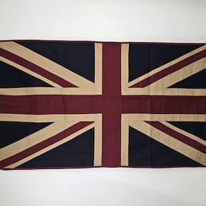 Pode incluir: Uma bandeira retangular com o design da Union Jack. A bandeira tem um fundo azul marinho com listras e formas vermelhas, bege e brancas. A bandeira tem uma borda bege com dois ilhós no lado esquerdo.