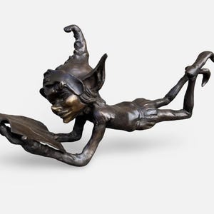 Könnte beinhalten: Eine Bronzeskulptur einer skurrilen Elfenfigur, die liegt und ein blattförmiges Objekt liest. Der Elf hat spitze Ohren, eine spitze Mütze und einen lächelnden Ausdruck. Die Skulptur ist detailliert mit einer dunkelbronzenen Oberfläche.