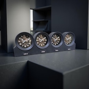 May include: A set of four black analog clocks displaying different time zones: Paris, London, Tokyo, and New York. Each clock has a dark face with visible gears and white hands. The clocks are mounted on a rectangular base.