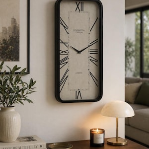 May include: A rectangular wall clock with a black frame and a cream face. The clock has black Roman numerals and hands, with the text "Kensington Station London". A table holds a lamp, candle, and decorative items. The clock measures approximately 60 cm in height.