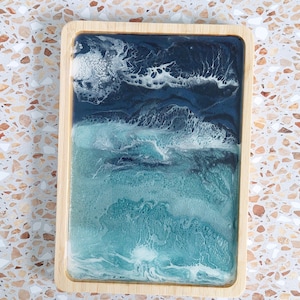 Peut inclure: Un plateau rectangulaire en bois avec une incrustation en résine imitant une scène océanique. Le motif passe d'un bleu marine profond en haut à un turquoise clair et un vert écume en bas, avec des motifs d'écume blanche dentelée.