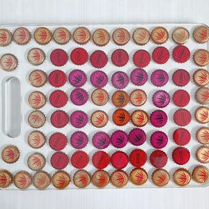 Peut inclure: Une planche à découper en acrylique transparente avec une poignée, décorée d'une mosaïque de capsules de bouteilles dans des tons de rouge, rose et or.