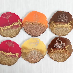 Pode incluir: Seis  tapetes redondos de resina com acabamento de glitter dourado e desenhos de folhas brancas. Os tapetes são em tons de vermelho, laranja e marrom.