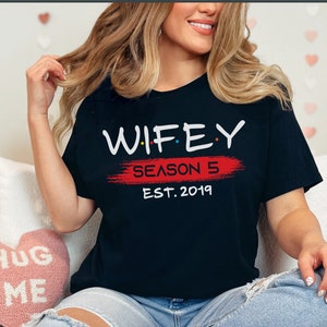 May include: A black t-shirt with white text that reads "WIFEY SEASON 5 EST. 2019" in a stylized font. The text is on a red brushstroke.