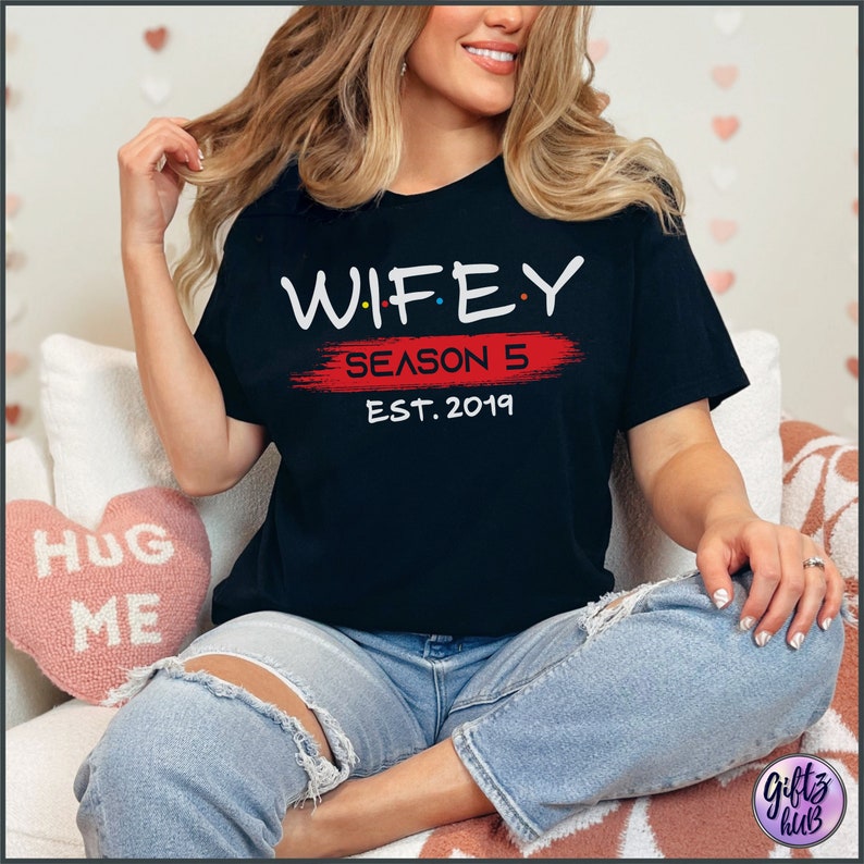 May include: A black t-shirt with white text that reads "WIFEY SEASON 5 EST. 2019" in a stylized font. The text is on a red brushstroke.