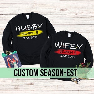 May include: Two black long-sleeved shirts with a white graphic print. The left shirt says "Hubby Season 5 Est. 2018" and the right shirt says "Wifey Season 5 Est. 2018".