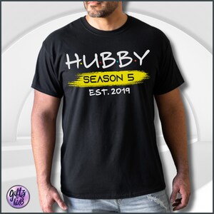 May include: Black t-shirt with white text that reads "HUBBY SEASON 5 EST. 2019". The text is in a stylized font and the word "SEASON" is written in yellow with a yellow paintbrush stroke behind it.