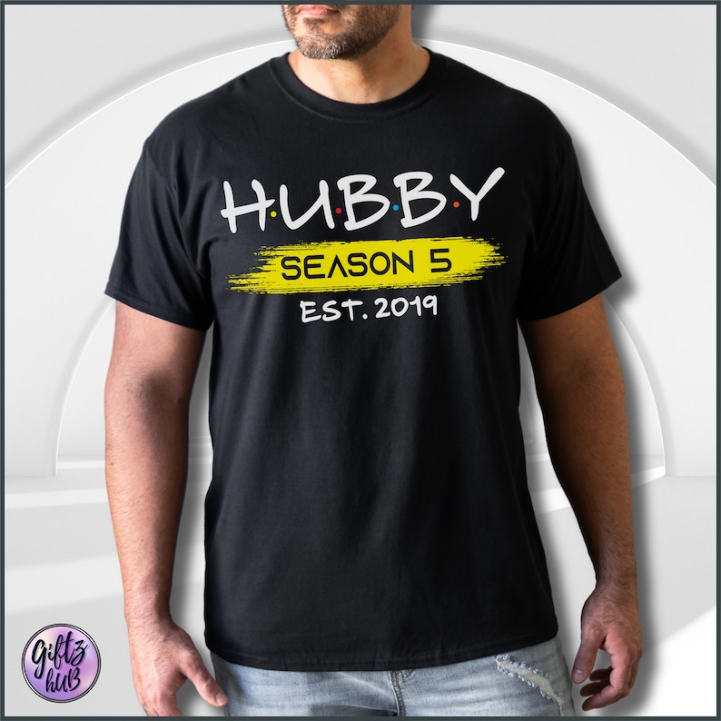 May include: Black t-shirt with white text that reads "HUBBY SEASON 5 EST. 2019". The text is in a stylized font and the word "SEASON" is written in yellow with a yellow paintbrush stroke behind it.