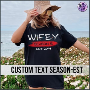 May include: Black t-shirt with the text "WIFEY SEASON 5 EST. 2019" in white and red. The text is stylized to look like the Friends logo.