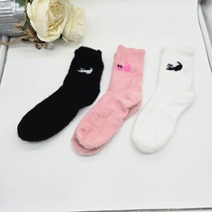 May include: Three pairs of fuzzy socks with different colors and designs. One pair is black with a white logo, one pair is pink with a pink logo, and one pair is white with a black logo.