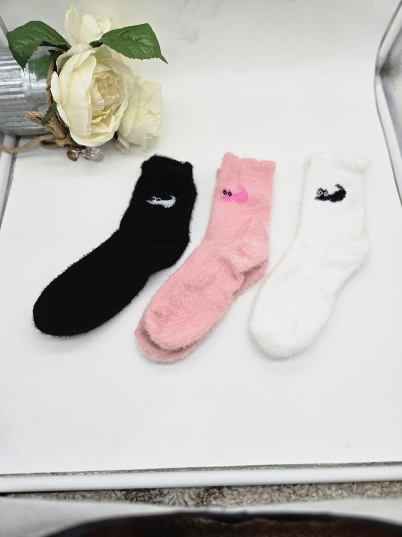 May include: Three pairs of fuzzy socks with different colors and designs. One pair is black with a white logo, one pair is pink with a pink logo, and one pair is white with a black logo.