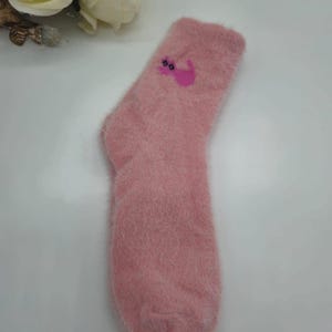 May include: A pair of pink fuzzy socks with a pink cat design. The cat has pink ears and a pink tail.