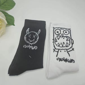 May include: A pair of black and white socks with a cartoon monster design. The black sock has a white cartoon monster with horns and the white sock has a black cartoon monster with a big mouth.