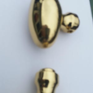 May include: Two gold-colored cabinet knobs. One knob is oval-shaped with a small, round protrusion. The other knob is a cylindrical shape with a rounded top. Both knobs have a shiny, reflective surface.