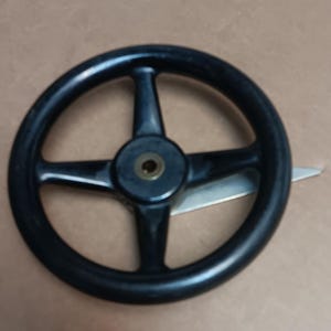 May include: A black circular handwheel featuring four spokes radiating from a central hub with a brass-colored metal insert. A silver-toned metal blade or lever extends from behind the wheel, visible on the right side of the central hub.