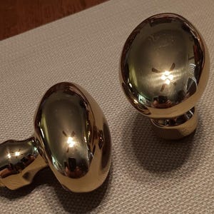 May include: Two polished gold-tone metal cabinet knobs with a smooth, rounded, egg-shaped head and a cylindrical base. The reflective surface shows light reflections and the surrounding environment, highlighting the metallic finish of the hardware.