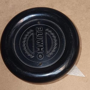 May include: A round, black Ohmite control knob featuring a central circular design with the word OHMITE embossed in the middle. The knob has a smooth, glossy finish and a small metal indicator pointer extending from the bottom right edge.