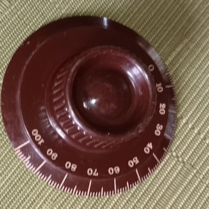 May include: A dark brown rotary dial with white markings, numbered from 0 to 100. The dial has a circular design with a raised center and a ridged edge. The numbers are evenly spaced around the perimeter.