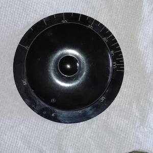 May include: A black, circular dial with white markings and numbers. The dial has a central knob and a series of graduated markings from 0 to 100. The letters "MC" are printed on the dial.